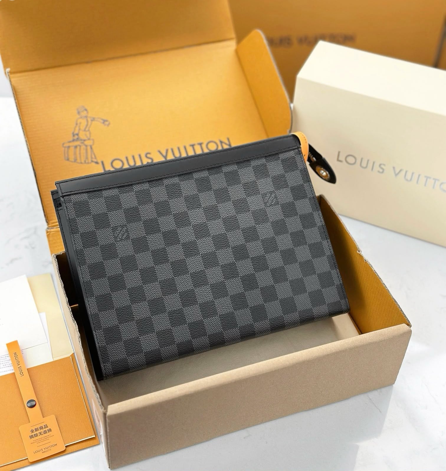 A Louis Vuitton zip pouch in the dark gray and black Damier Graphite canvas, displayed inside its open cardboard packaging box on a white marble surface.