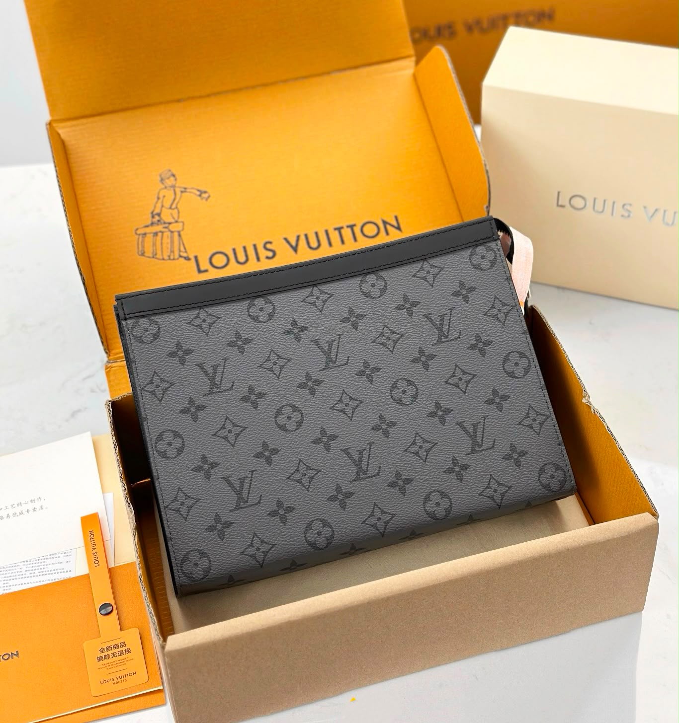 A Louis Vuitton monogram pouch in dark gray coated canvas, displayed inside its orange Louis Vuitton box alongside branded packaging and a cream-colored box.