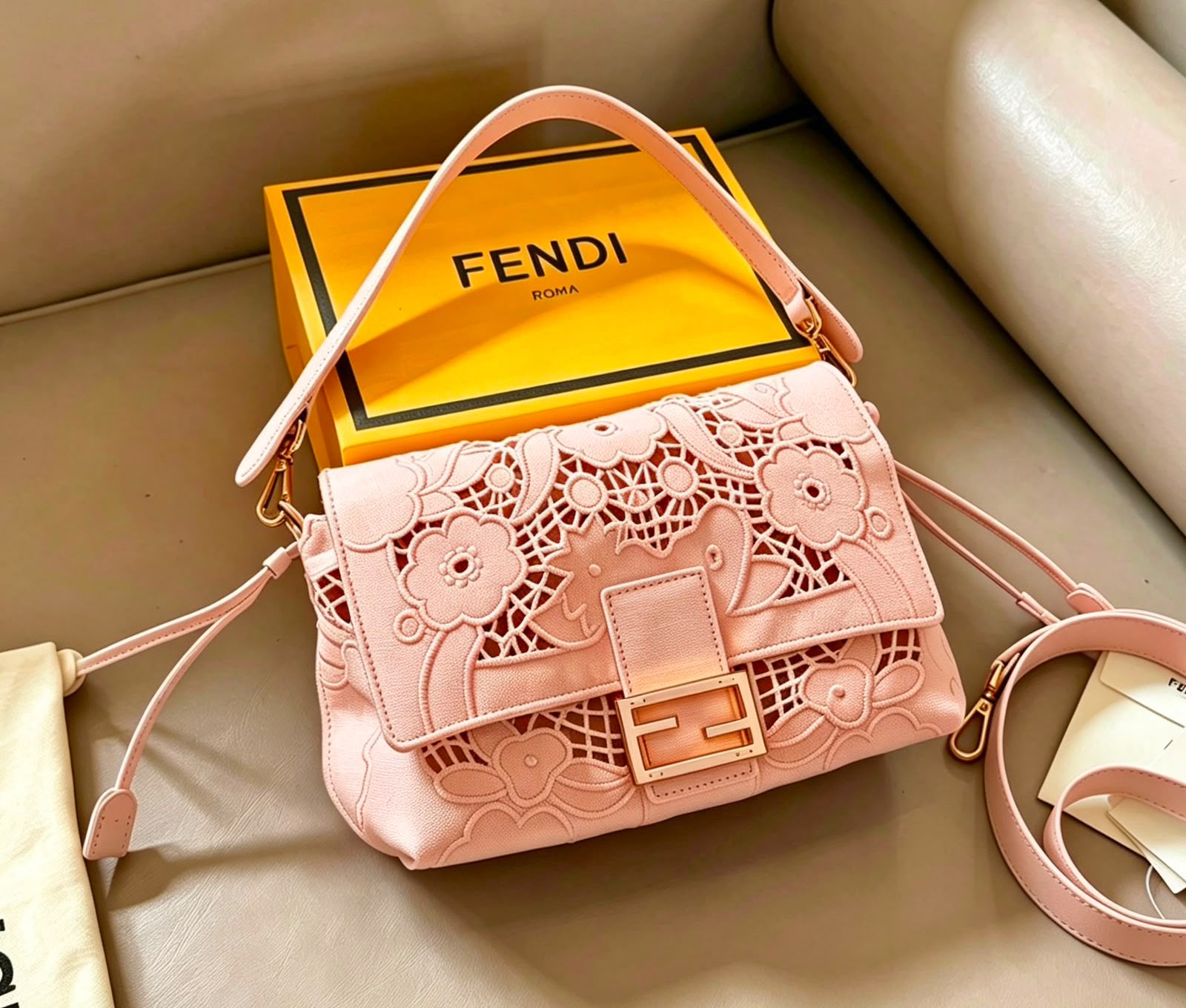 A pink Fendi shoulder bag with floral laser-cut detailing on the front flap, displayed next to its signature yellow Fendi Roma box on a beige leather surface.
