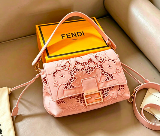 A pink Fendi shoulder bag with floral laser-cut detailing on the front flap, displayed next to its signature yellow Fendi Roma box on a beige leather surface.