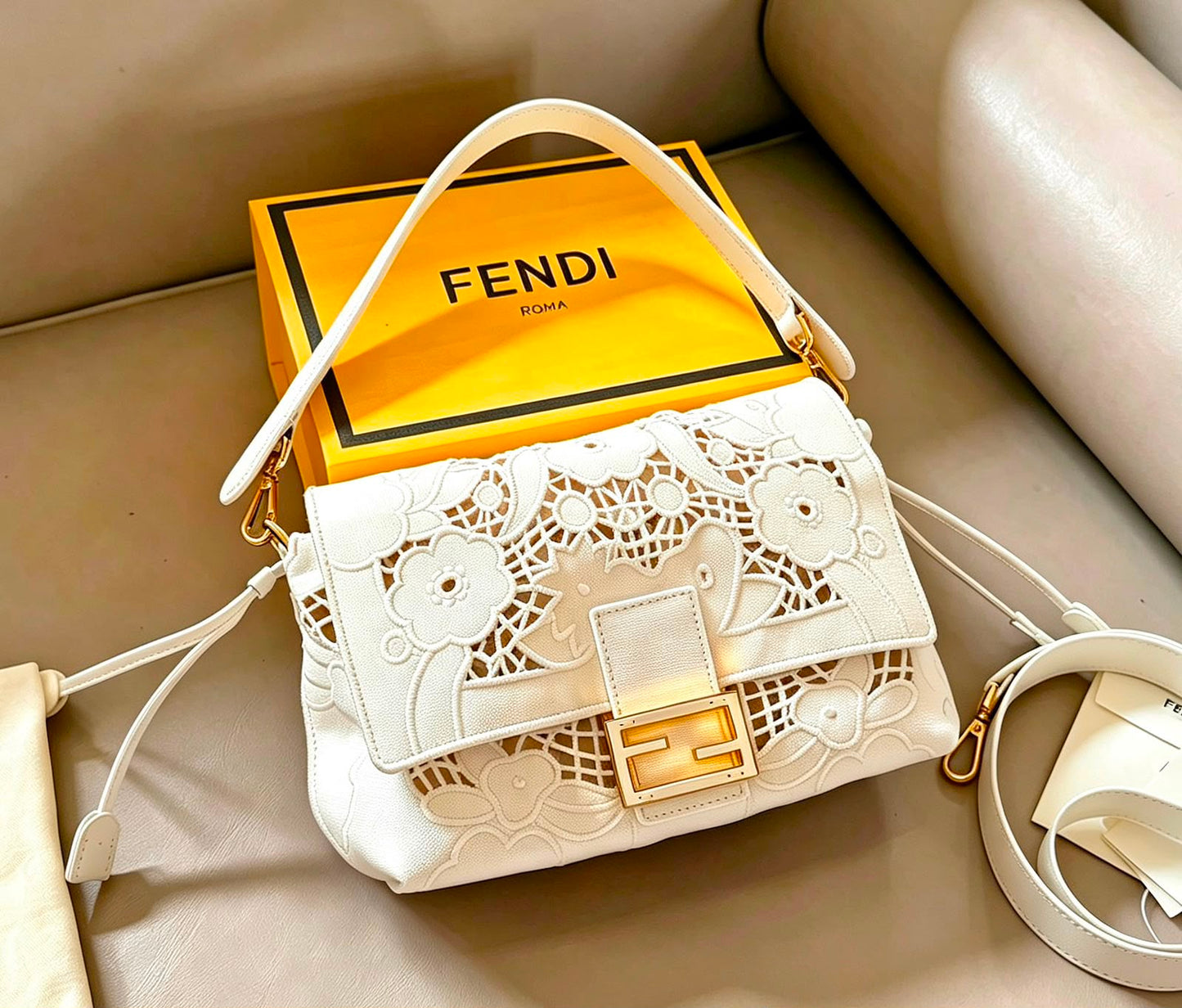 A white Fendi shoulder bag with floral cutout detailing and a gold FF clasp, displayed on a beige leather surface with its signature yellow Fendi box behind it.