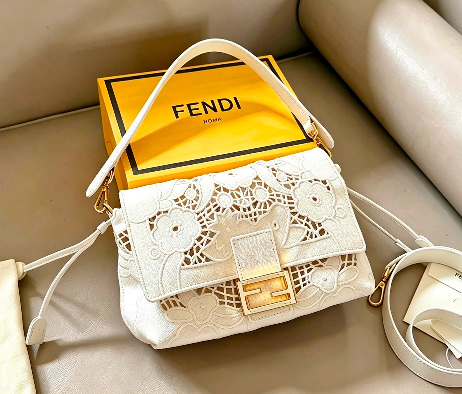 A white Fendi shoulder bag with floral cutout detailing and a gold FF clasp, displayed on a beige leather surface with its signature yellow Fendi box behind it.