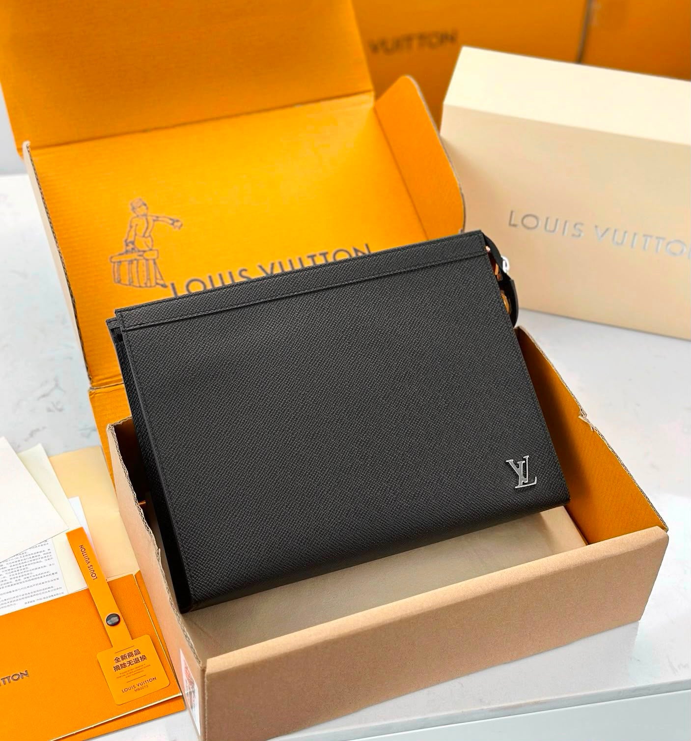 A Louis Vuitton black leather pouch with a silver-tone LV logo, displayed inside its open orange and beige packaging boxes.